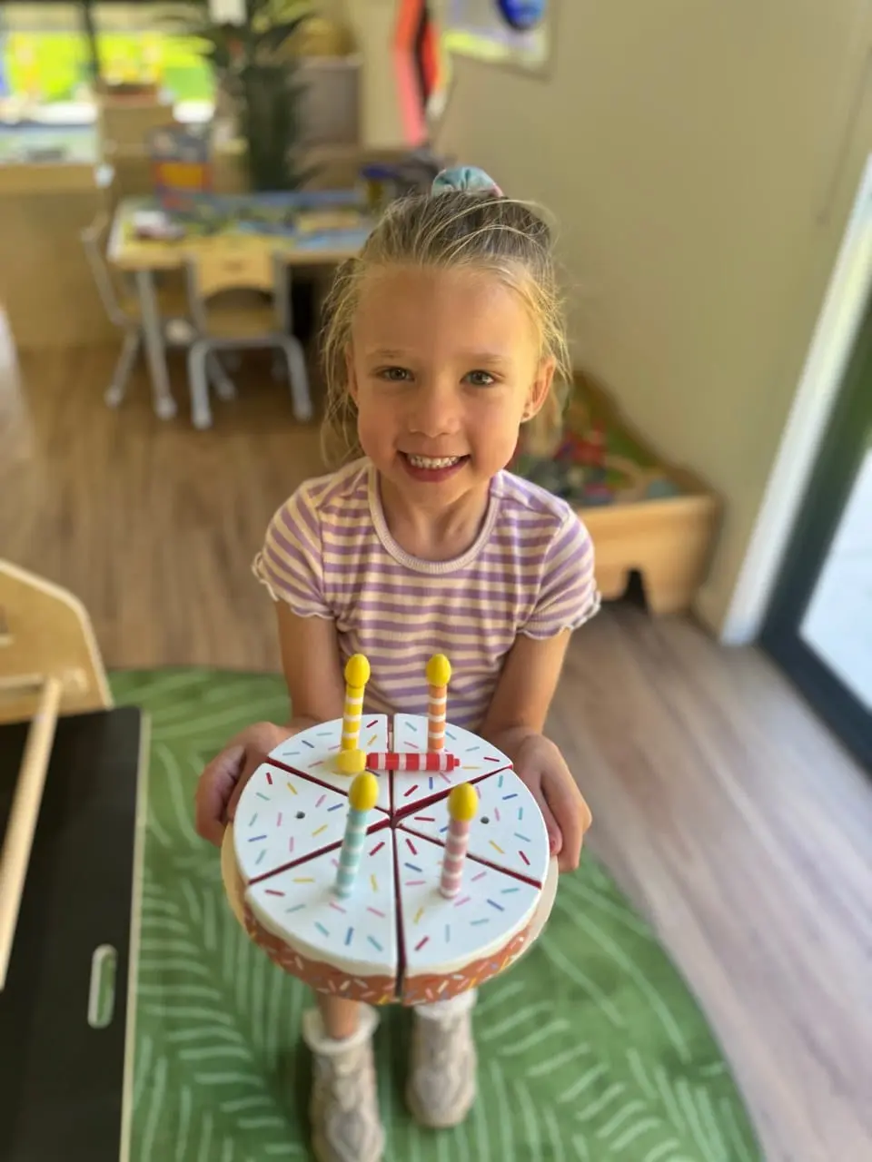 A child proudly showing off a wooden birthday cake during imaginative play at Kids Kingdom.